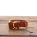 The Leather Dog Collar by Ballad of the Bird Dog is handmade in Brenham from brown English bridle leather, features a brass buckle and D-ring, and is logo-stamped—shown resting on a wooden surface.