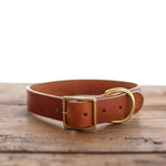 The Ballad of the Bird Dog Leather Dog Collar, crafted from brown leather with a brass buckle and D-ring, is showcased on wood for a classic look against a clean white background.