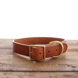 The Leather Dog Collar by Ballad of the Bird Dog features handmade brown leather, a brass buckle, and D-ring, shown on wood with a white background. Crafted in Brenham for timeless quality and style—perfect for your dog.