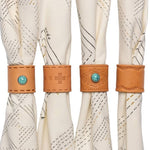 Four cream-colored fabric napkins are each held by a Jenni Earle Leather Studded Bandana Slide—three decorated with turquoise stones and stitching, and one with plain stitching—for a handcrafted, Western-inspired table setting.