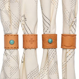 Four cream-colored fabric napkins are each held by a Jenni Earle Leather Studded Bandana Slide—three decorated with turquoise stones and stitching, and one with plain stitching—for a handcrafted, Western-inspired table setting.