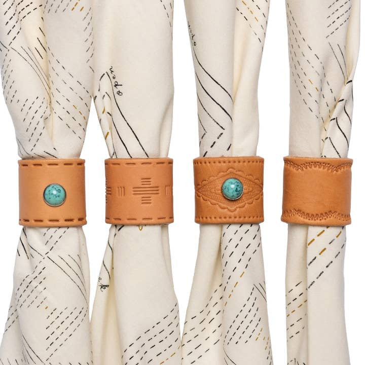 Four cream-colored fabric napkins are each held by a Jenni Earle Leather Studded Bandana Slide—three decorated with turquoise stones and stitching, and one with plain stitching—for a handcrafted, Western-inspired table setting.
