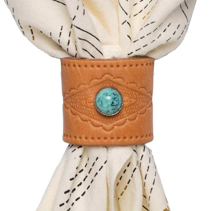 The Jenni Earle Leather Studded Bandana Slide features brown genuine oiled leather with a turquoise stone accent and holds a cream napkin with black stitched lines.