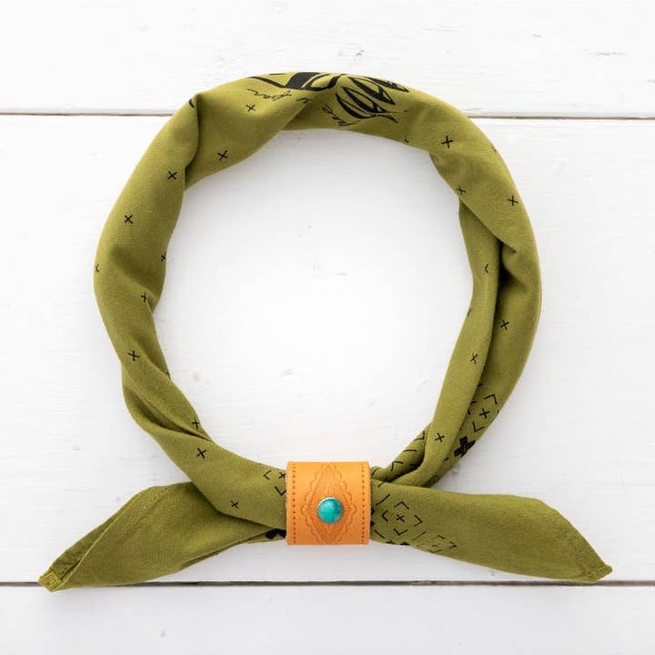 The Jenni Earle Leather Studded Bandana Slide secures a green bandana with black designs, featuring genuine oiled leather and a turquoise button, shown on a white wooden surface.