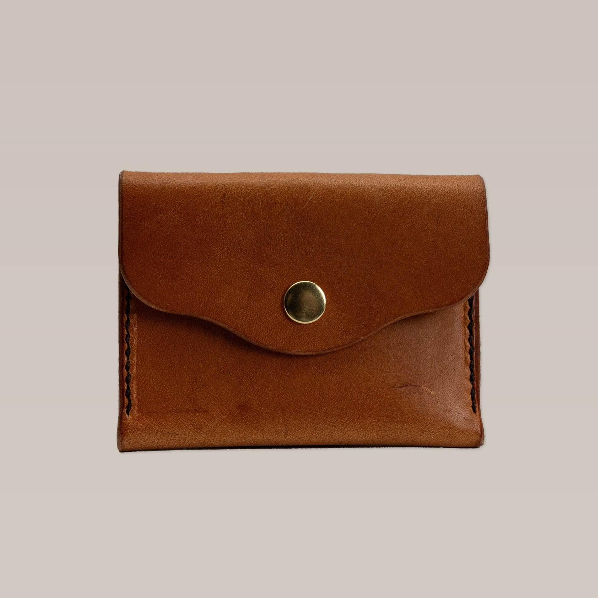 The Leather Wallet by Ballad of the Bird Dog is a small card wallet in brown leather with a flap and gold button snap closure, shown on a plain beige background.