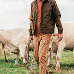 A man wearing lightweight Duck Camp Brush Pants and a cap holds a coiled rope while walking in a field among white cows.