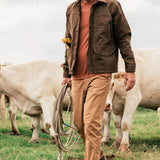 A man wearing lightweight Duck Camp Brush Pants and a cap holds a coiled rope while walking in a field among white cows.