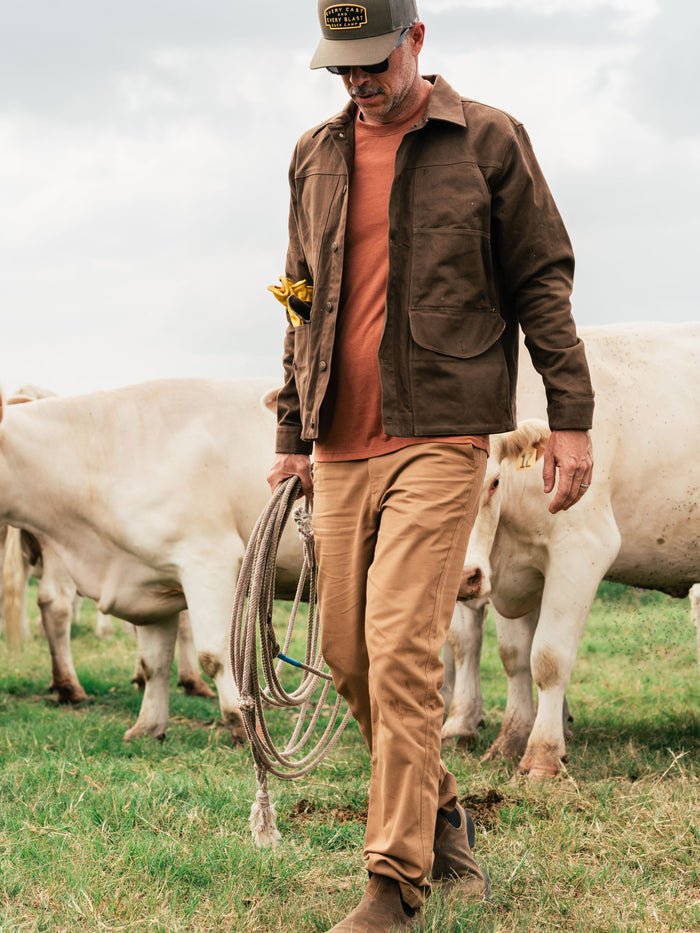 A man wearing lightweight Duck Camp Brush Pants and a cap holds a coiled rope while walking in a field among white cows.