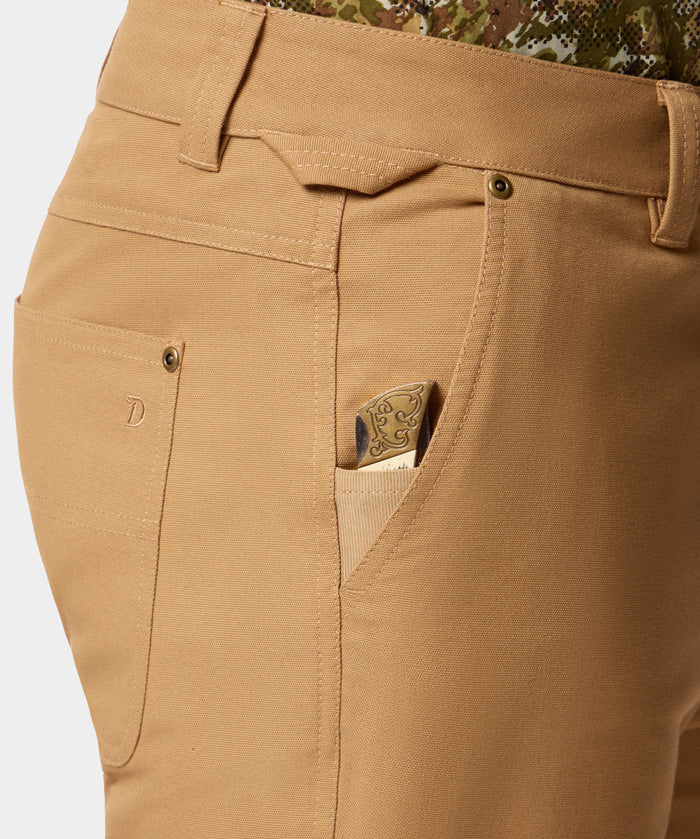 Close-up of Duck Camp Lightweight Brush Pants, featuring a durable cotton blend and a patterned pocket knife partially visible in the front coin pocket; worn with a green and brown camouflage shirt.