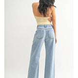 A woman stands with her back to the camera, wearing the JBD Lightweight Drapey Relaxed Trouser, wide-leg light denim jeans, paired with a strapless beige top against a plain white background.