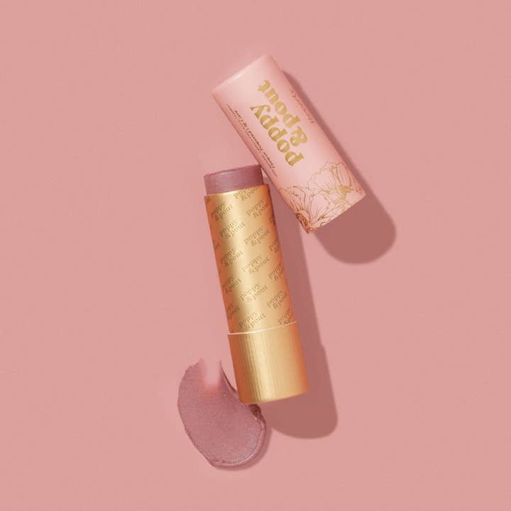A tube of Poppy & Pout's Lip Balm with the cap off reveals a pinkish tint, alongside a swatch of the cruelty-free balm smeared on a pink surface.