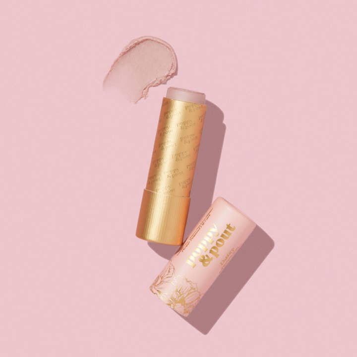 A gold and pink tube of Poppy & Pout Lip Balm lies open on a pink background with a smear of balm above it.