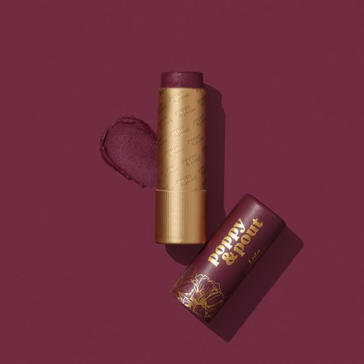 A cruelty-free Poppy & Pout Lip Balm with a gold tube and matching burgundy cap labeled “poppy & pout” sits on a solid burgundy background.