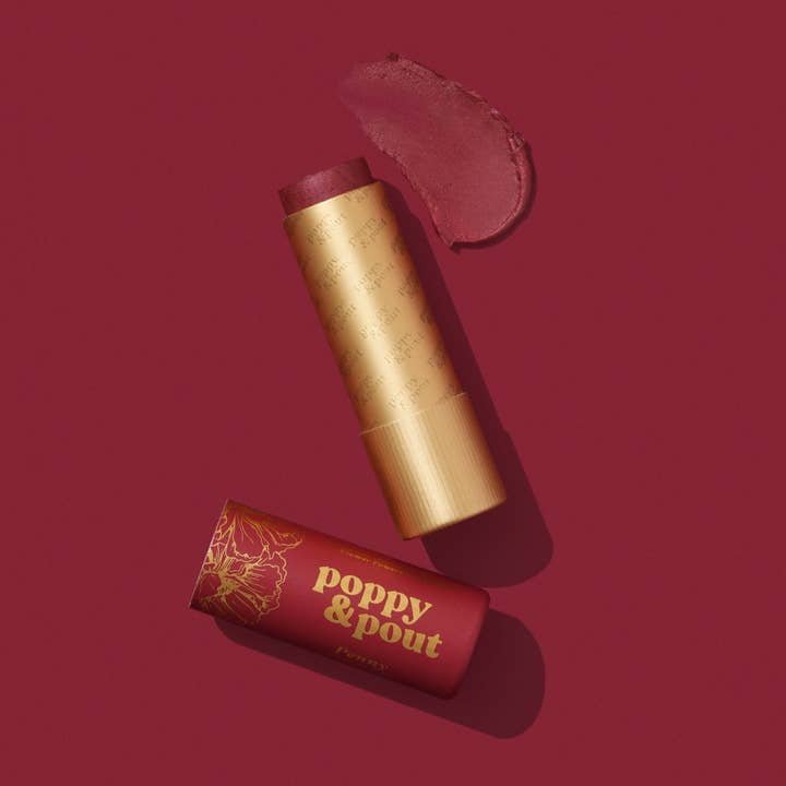 A tube of Poppy & Pout’s Lip Balm with the cap off displays the hand-poured product and a swatch on a matching red background.