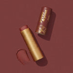A tube of Poppy & Pout Lip Balm, shown uncapped with gold lettering and a cruelty-free swatch on a brown background, highlights the brand’s signature color.