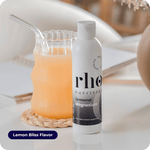 A bottle of Liposomal Magnesium | Rho Nutrition sits by a glass of orange drink with a glass straw on a white table. The "Lemon Bliss Flavor" label highlights magnesium for muscle recovery and nervous system support.