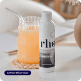 A bottle of Liposomal Magnesium | Rho Nutrition sits by a glass of orange drink with a glass straw on a white table. The "Lemon Bliss Flavor" label highlights magnesium for muscle recovery and nervous system support.