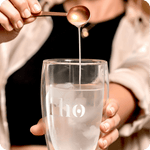 A person pours liquid from a spoon into an ice water glass labeled "rho"—an everyday ritual with Liposomal NAD+ by Rho Nutrition, designed to support healthy aging and cellular energy.