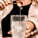 A person holds a water glass with ice, pouring a liquid from a spoon into it. The Liposomal NAD+ by Rho Nutrition suggests support for cellular energy and healthy aging through NAD+ supplementation.