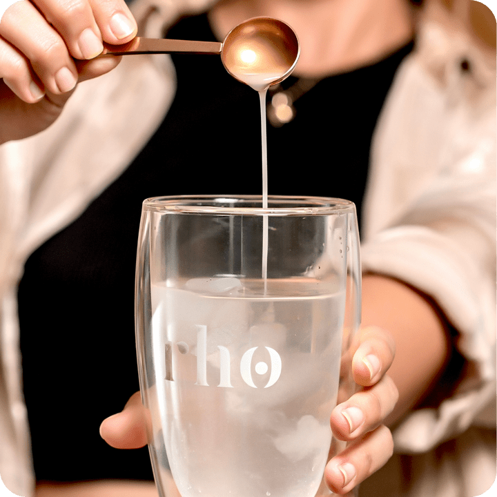 A person holds a water glass with ice, pouring a liquid from a spoon into it. The Liposomal NAD+ by Rho Nutrition suggests support for cellular energy and healthy aging through NAD+ supplementation.