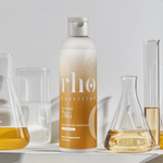 A bottle of Liposomal TMG by Rho Nutrition sits on a white surface, with three glass beakers filled with yellow liquids in the background, highlighting support for healthy homocysteine metabolism.