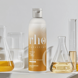 A bottle of Liposomal TMG by Rho Nutrition sits on a white surface, with three glass beakers filled with yellow liquids in the background, highlighting support for healthy homocysteine metabolism.