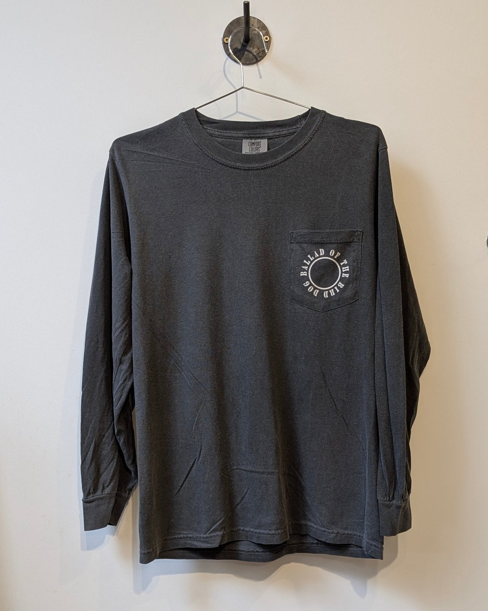 The Shop Shirt Long Sleeve | Thunderbird by Ballad of the Bird Dog features a dark gray design with a chest pocket, white circular eye graphic, and "OVAL OF GOD WEAR," making it your perfect adventure shirt.