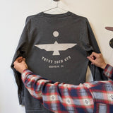 A person holds the Ballad of the Bird Dog Shop Shirt Long Sleeve | Thunderbird, featuring a geometric bird and "TRUST YOUR GUT, BRENHAM, TX" on the back—an adventure-ready top for explorers, seen hanging on a wall hook.
