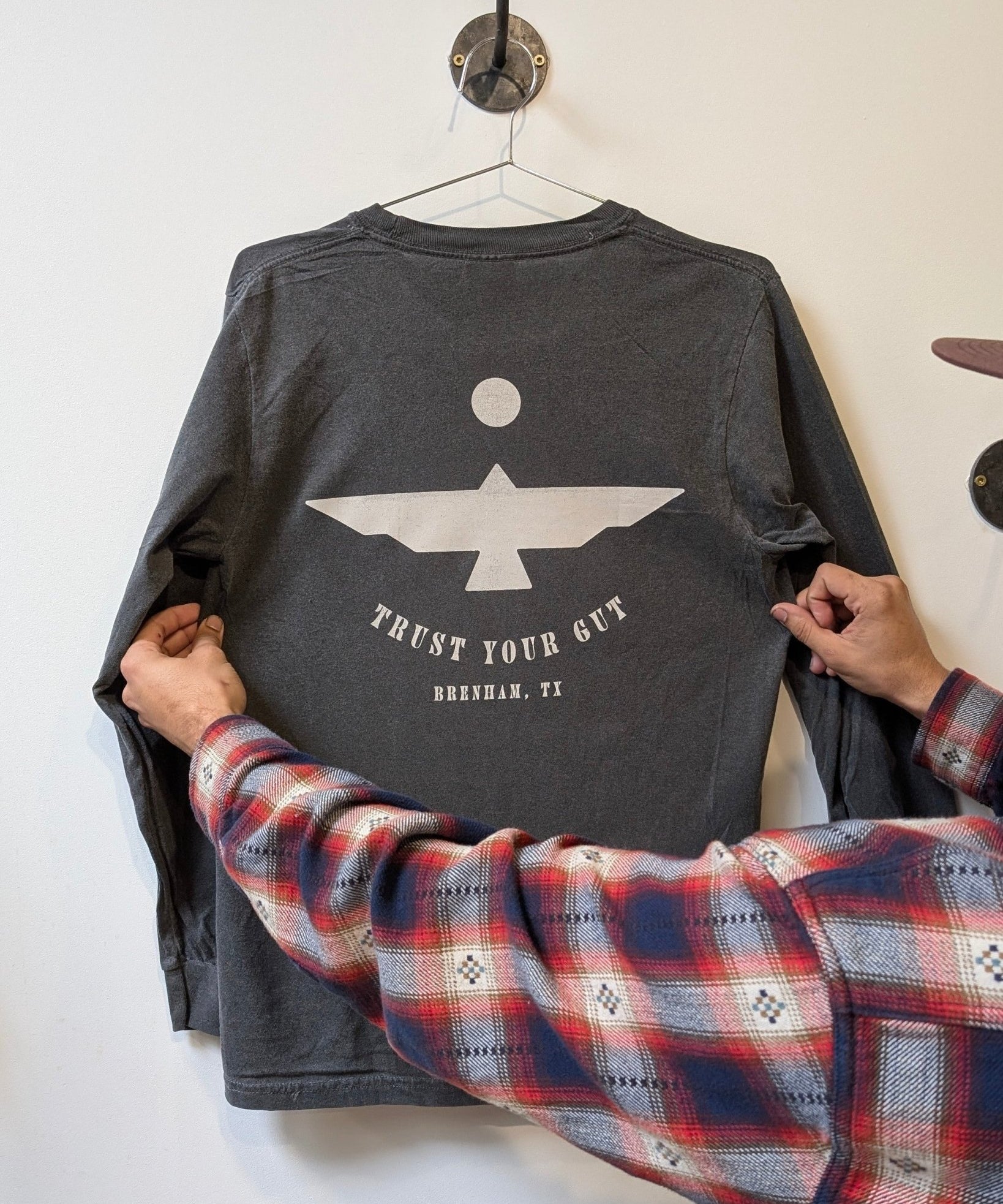 A person holds the Ballad of the Bird Dog Shop Shirt Long Sleeve | Thunderbird, featuring a geometric bird and "TRUST YOUR GUT, BRENHAM, TX" on the back—an adventure-ready top for explorers, seen hanging on a wall hook.