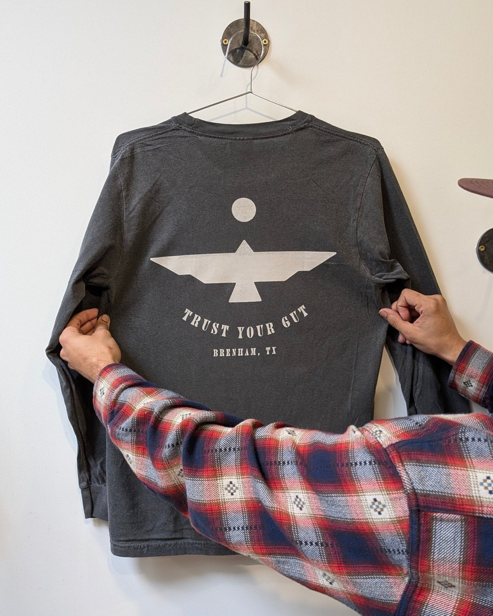 A person holds the Ballad of the Bird Dog Shop Shirt Long Sleeve | Thunderbird, featuring a geometric bird and "TRUST YOUR GUT, BRENHAM, TX" on the back—an adventure-ready top for explorers, seen hanging on a wall hook.