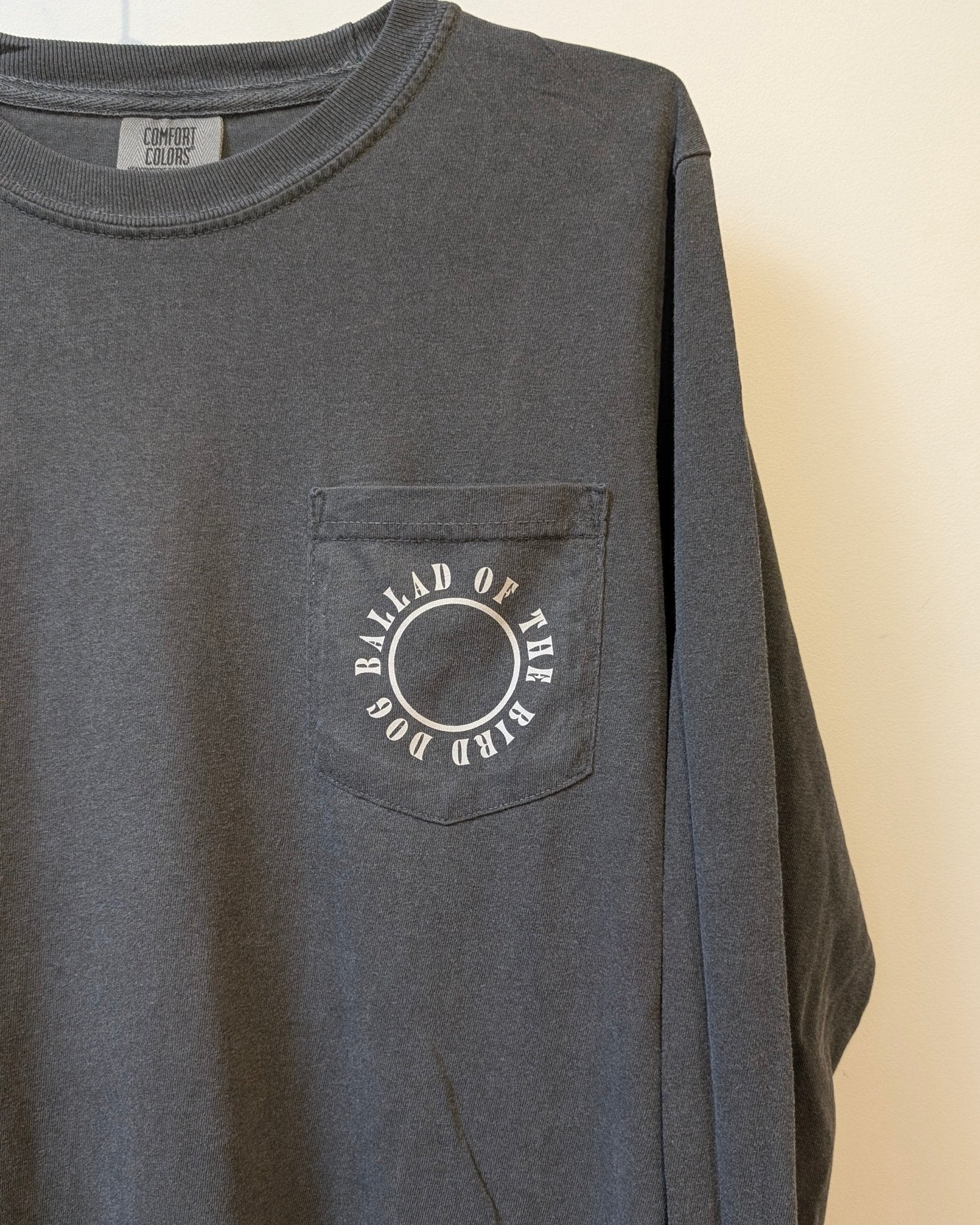 A dark gray Shop Shirt Long Sleeve | Thunderbird by Ballad of the Bird Dog featuring a chest pocket with a circle and "BALLAD OF THE BIRD DOG" printed around it.