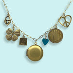 The Ornamental Things Love and Luck Charm Necklace showcases a gold cable chain with a locket, four-leaf clover, “LOVE” square, blue heart, “AMOUR” circle, peace sign, and heart charms on a light blue background.