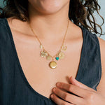 A woman wears the Ornamental Things Love and Luck Charm Necklace, featuring gold charms—a flower, heart, round locket, and more—on a gold cable chain over a dark sleeveless top.