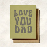 The "Love You Dad" Birthday Card by Daydream Prints sits on a sandy surface, showcasing bold gray letters—an ideal choice to celebrate Father's Day.