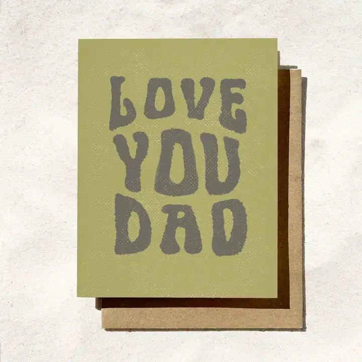 The "Love You Dad" Birthday Card by Daydream Prints sits on a sandy surface, showcasing bold gray letters—an ideal choice to celebrate Father's Day.