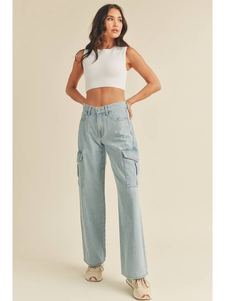 A woman stands with hands on hips against a plain background, wearing a white sleeveless crop top, JBD's Lt - Cargo Jean 7-pocket high-rise jeans in light blue, and beige sneakers.