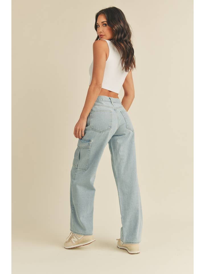 A woman faces away, wearing a white sleeveless crop top, JBD Lt - Cargo Jean high-rise light blue jeans with side pockets, and beige sneakers against a plain background.