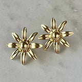 The Lucienne Earrings by Jessica Matrasko Jewelry feature delicate gold filled daisy studs with petal designs, displayed on light marble. These lightweight, hypoallergenic earrings are perfect for everyday elegance.