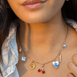 A woman wears the Peter and June Lucky Charmed Necklace—an 18k gold plated, waterproof piece featuring assorted charms like a fish, cherries, heart, and ceramic-like pendants, all made from ethically sourced materials.
