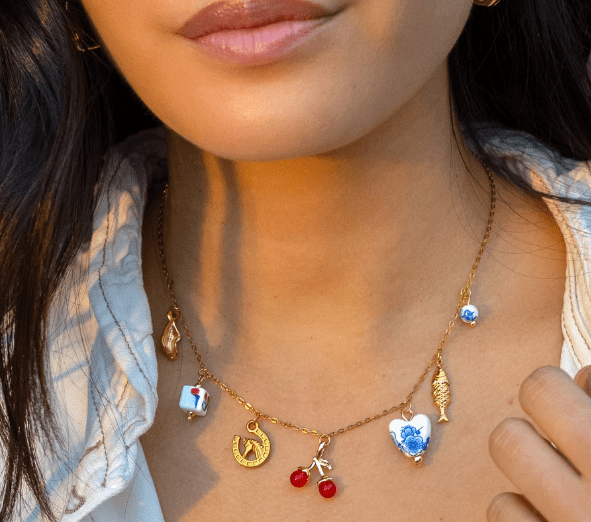 A woman wears the Peter and June Lucky Charmed Necklace—an 18k gold plated, waterproof piece featuring assorted charms like a fish, cherries, heart, and ceramic-like pendants, all made from ethically sourced materials.