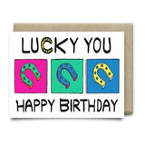 The "Lucky You Happy Birthday Card" by Anvil Cards is A2-sized, blank inside, and features three colored horseshoes in pink, green, and blue squares on elegant linen paper with "Lucky You" above and "Happy Birthday" below.