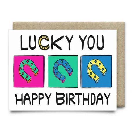 The "Lucky You Happy Birthday Card" by Anvil Cards is A2-sized, blank inside, and features three colored horseshoes in pink, green, and blue squares on elegant linen paper with "Lucky You" above and "Happy Birthday" below.