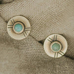 The Madison Studs by CommonForm feature two round brass discs with central turquoise stones and engraved line patterns, displayed on textured beige fabric.