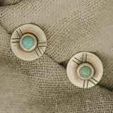 The Madison Studs by CommonForm feature round brushed brass discs with turquoise centers and engraved lines, displayed on beige textured fabric.