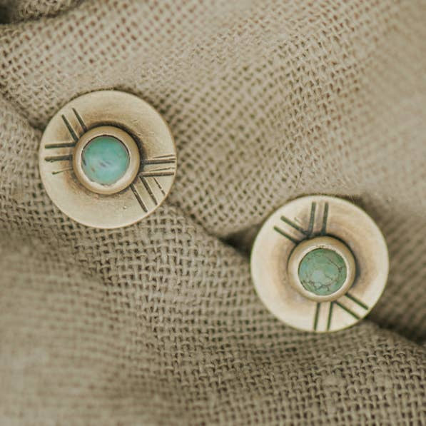 The Madison Studs by CommonForm feature round brushed brass discs with turquoise centers and engraved lines, displayed on beige textured fabric.