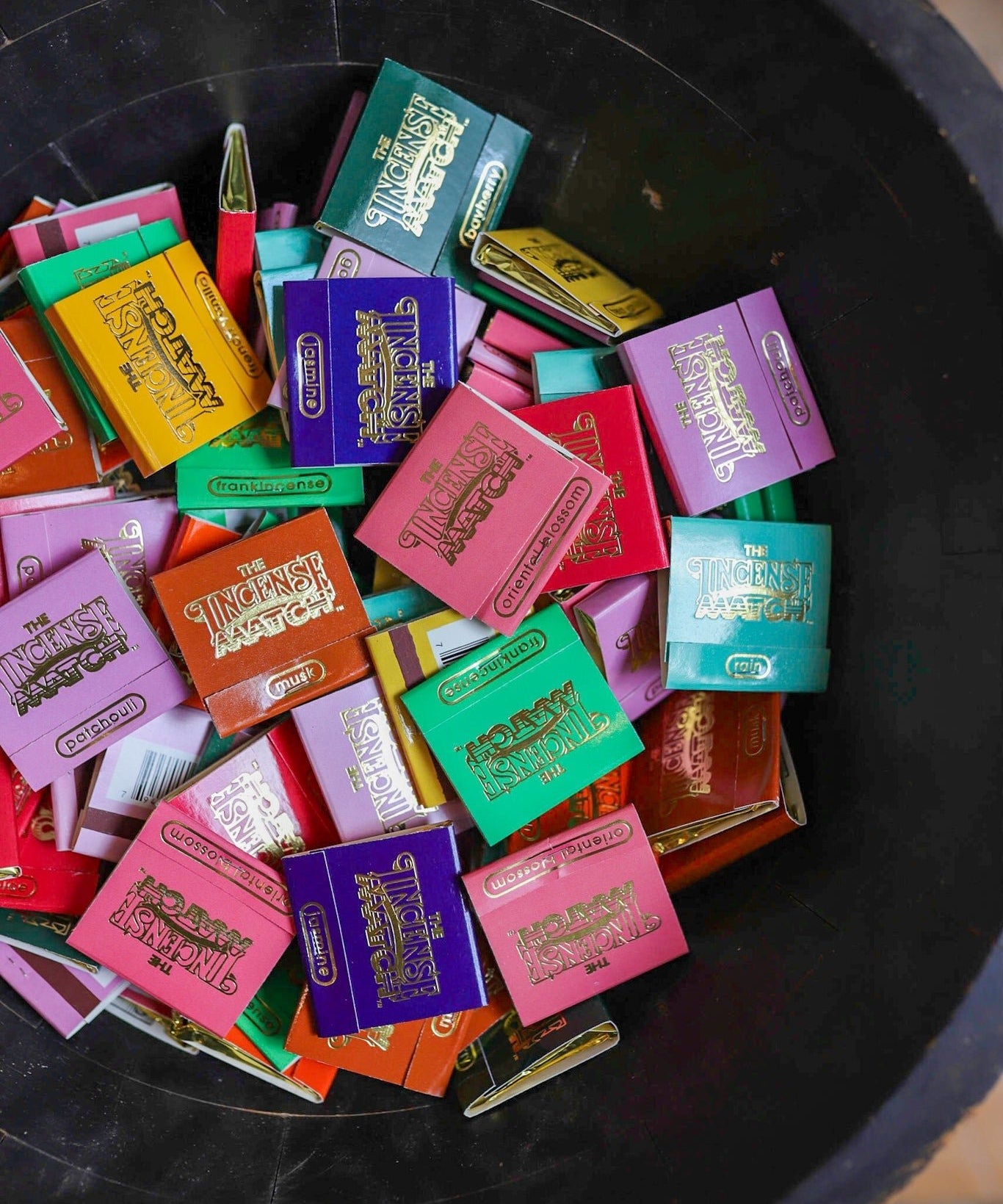 A black container from The Incense Match holds colorful matchbook-style packets labeled "The Strongest Matcha," perfect for storing incense matches or using as a unique air freshener.