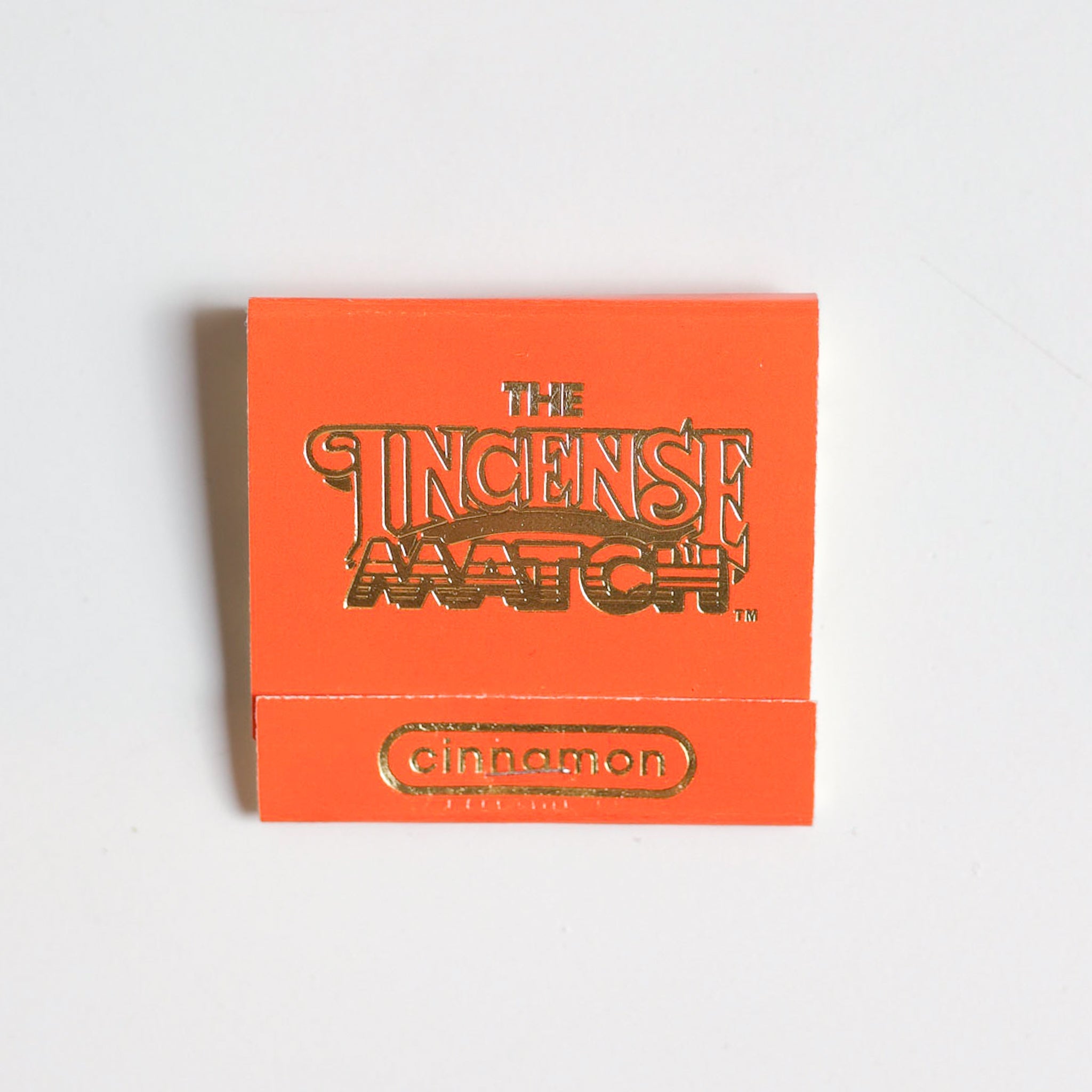 An orange matchbook labeled "Matches | The Incense Match" by The Incense Match with "cinnamon" in gold lettering rests on a white surface—perfect as a compact air freshener and thoughtful incense gift.