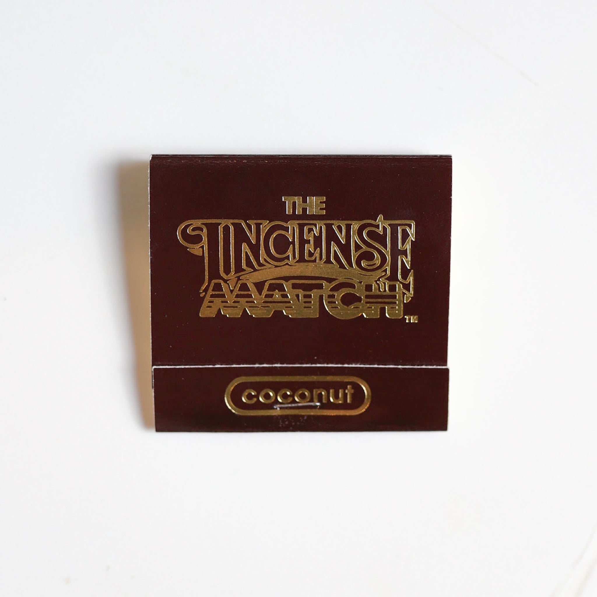 A brown matchbook labeled "Matches | The Incense Match" by The Incense Match with "coconut" at the bottom rests on a white surface—ideal for enjoying incense matches that also freshen the air with coconut fragrance.