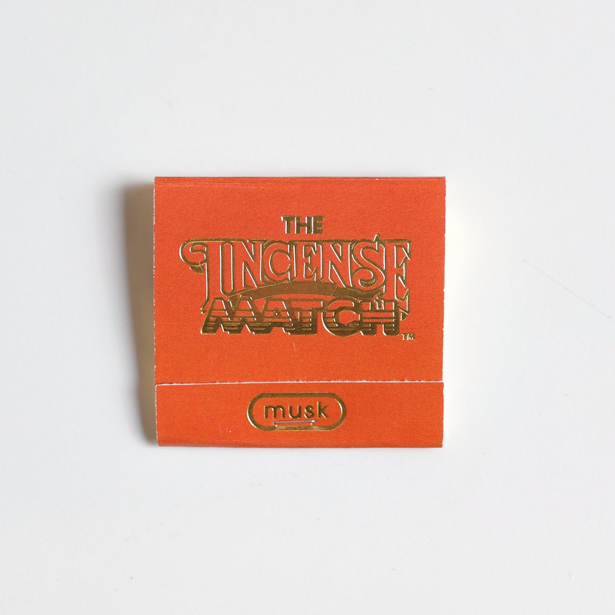 An orange "Matches | The Incense Match" booklet from The Incense Match brand, labeled "musk," sits on a white background—ideal as an incense gift or air freshener with convenient incense matches.