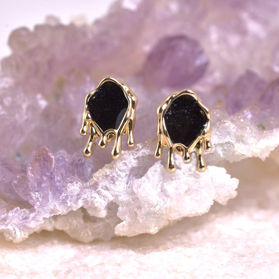 The Melting Gem Gold Stud Earrings by cameoko feature black stone accents and are displayed on a cluster of light purple and white crystals.